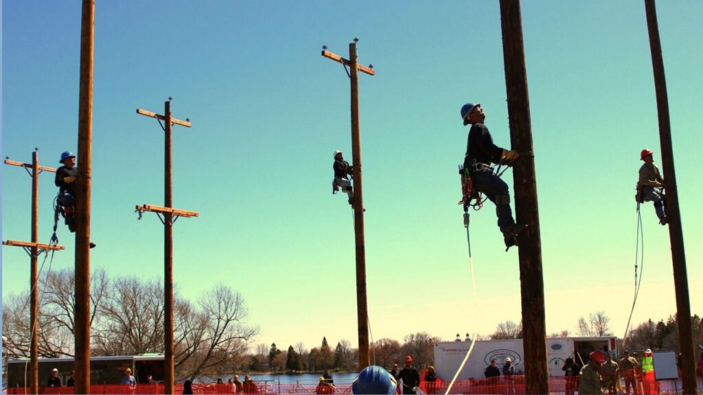 Lineman trainees climbing utility poles during an outdoor training event under a bright blue sky.